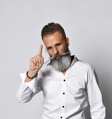 Portrait of a stylish, handsome and smart hipster man who raises his finger up on a white background. Middle aged man with a gray beard and mustache is wearing a white shirt. Thought concept.