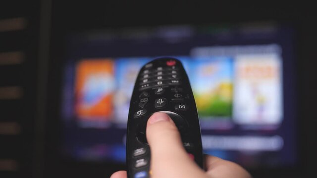 Man's hand selects internet tv channels with remote control, close-up. Person controls TV using a modern remote control. A man watches smart TV and uses black remote control. Blurry tv scrolls pages