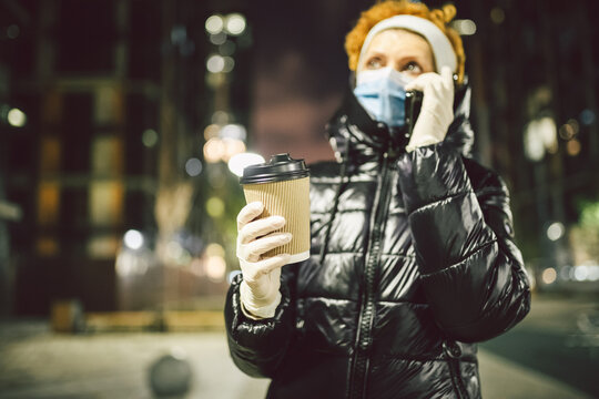 Adult woman is wearing covid protection mask and medical gloves as she walks along a European city during quarantine, drinks beverage and uses a cell phone. Theme coronavirus and life of citizens