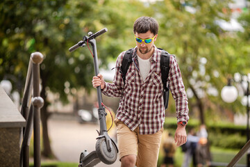 Modern city dweller caucasian man carries electric scooter over obstacle in park while driving around the city. Theme of convenient functional public space, city infrastructure and inclusiveness