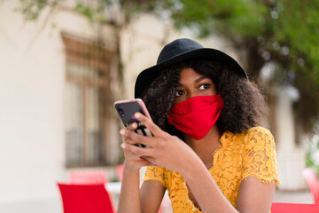 young black woman with curly hair, with red mask, yellow dress and black hat, checking her phone