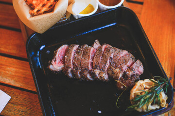Beef steak with mustard sauce and cucumber and pickles and basket of french fries placed on a wooden tray close up.