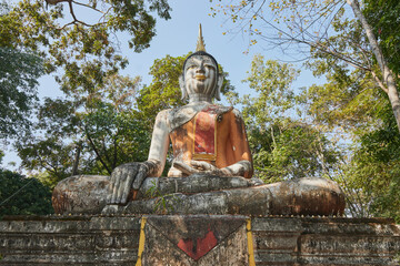 Fototapeta premium Phayao, Thailand - Dec 6, 2020: Low Angle Front Meditation Buddha Statue in Green Forest and Blue Sky Background in Wat Analayo Temple