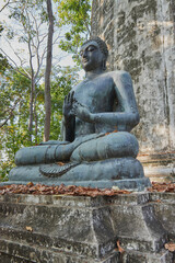 Fototapeta na wymiar Phayao, Thailand - Dec 6, 2020: Portrait Low Angle Front Left Buddha Statue Giving The Firts Sermon on Green Forest Background in Wat Analayo Temple