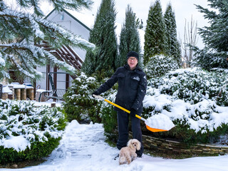 A 61-year-old European man with his dog is clearing snow with an orange shovel. Winter old age lifestyle concept