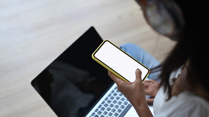 Close up view young female using mock up smart phone and laptop with blank screen while sitting on wooden floor.