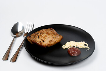 Tilted side view Black pepper pork steak, garnished with treetops and two dipping sauces, mayonnaise and tomato.On a round black plate. Metal spoons and forks placed on the sides. isolated background.