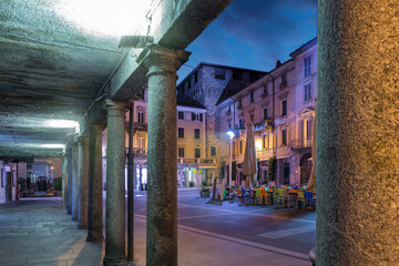Downtown deserted city street scene at night. Lecco, Italy. Picturesque town overlooking lake Como. Square XX Settembre and the old Viscontea tower seen from the arcades - obrazy, fototapety, plakaty
