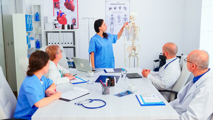Fototapeta premium Nurse giving presentation in front of doctors team about human anatomy using skeleton. Clinic expert therapist talking with colleagues about disease, medicine professional.