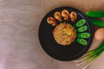 Shrimp fried rice in a black plate with spring onion, egg and cucumber all on the wooden table. top view.