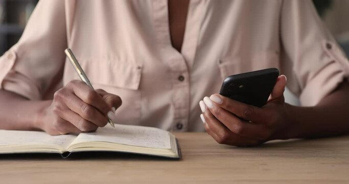 Mixed-race woman working sit at table hold cellphone writes schedule in diary with pen, close up. Student girl learns using internet resources. Create planning to-do list in personal organizer concept