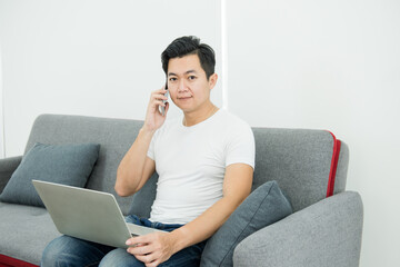 Happy asian man talking on smartphone while sitting on sofa and using laptop