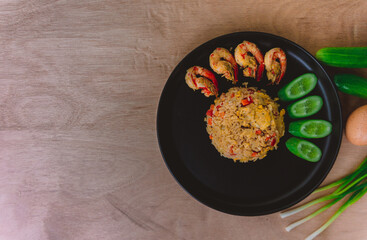 Shrimp fried rice in a black plate with spring onion, egg and cucumber all on the wooden table. top view.
