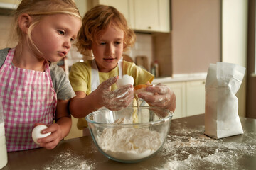 Two cute little children, boy and girl in aprons adding eggs while preparing dough together on the kitchen table at home