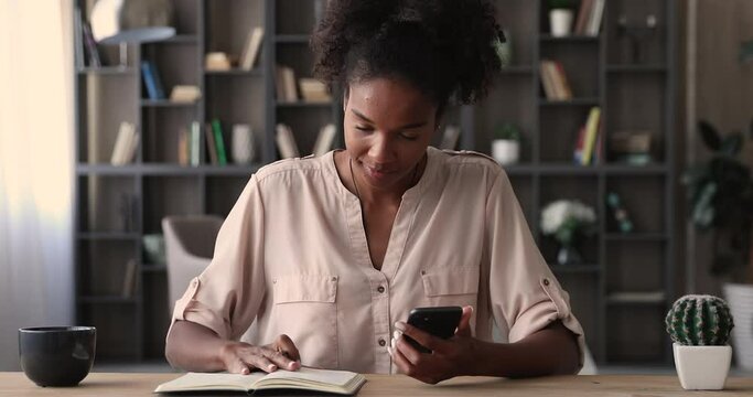 African woman sit at office desk holding smartphone writing event agenda on diary, businesswoman planning daily business appointment, create schedule plan in calendar, make list to-do reminder concept