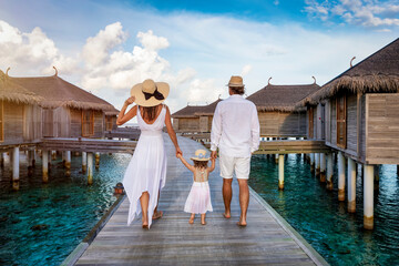 A family in white summer clothes walks over a wooden pier between over water lodges in the Maldives...