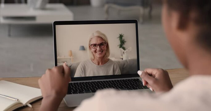 50s woman talk to colleague using video conference, laptop screen view over african female shoulder sit at desk take part in remote communication with elderly friend living abroad. Video call concept