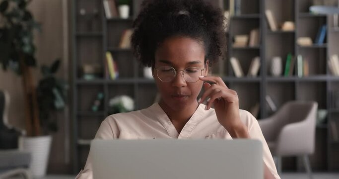 Pensive serious young african woman wear glasses lost in thoughts, working on online project, pondering strategy, searching solution and creative ideas, thinking over task sitting at desk with laptop