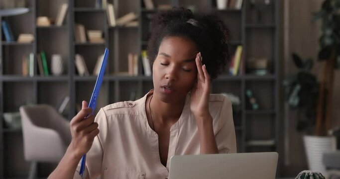 Young African jaded woman dying of heat in office without air conditioner at hot summer working day. Mixed race female sit at desk waving blue handheld fan relieving discomfort, hot temperature indoor