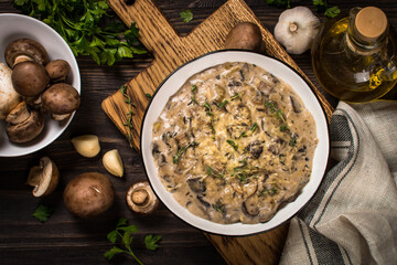 Mushrooms in cream sauce with herbs and cheese. Top view at dark wooden table with ingredients for cooking.