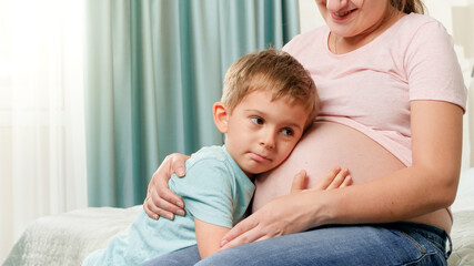 Portrait of little toddler boy hugging and listening to future baby in pregnant mothers belly
