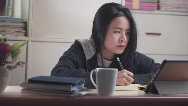 young asian female study from home by watching tablet online e-learning class lesson, hand writing with pencil on notebook, wear sweater warm night, distanced leaner, home school