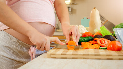 Closeup of pregnant woman with big belly cutting vegetables and cooking sald on kitchen.