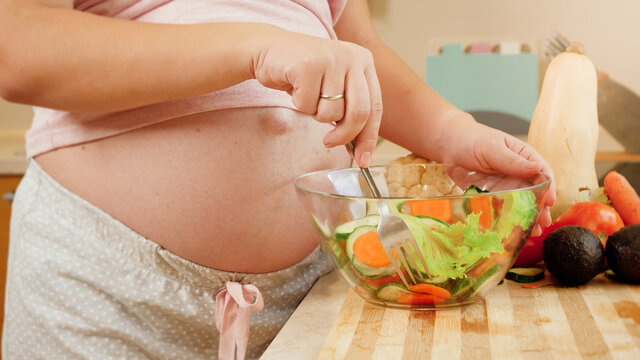 Closeup of yougn pregnant woman with big belly eating fresh vegetable salad while standing on kitchen. Concept of healthy lifestyle and nutrition during pregnancy