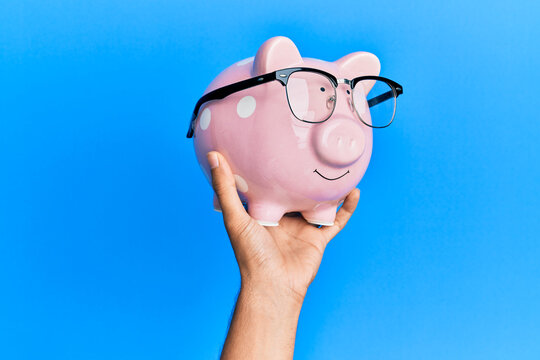 Hand Of Hispanic Man Holding Funny Piggy Bank With Glasses Over Isolated Blue Background.