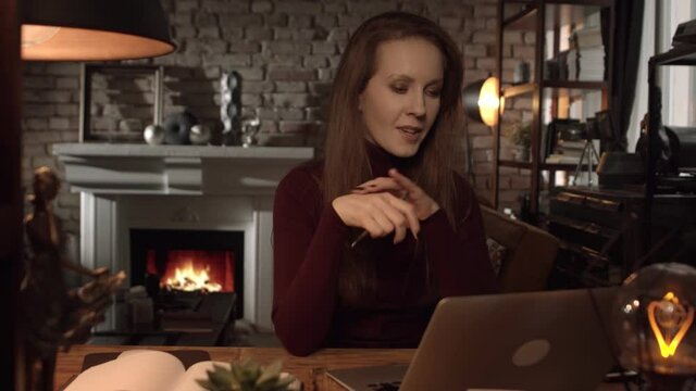 Home office - woman working from home with laptop computer. Talking on video chat, sitting at desk in dark living room in front of fireplace in Winter.