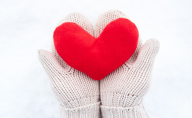 Creative greeting card for Valentines Day. Hold red soft heart toy made of fabric in beige mittens against background of white freshly fallen snow.