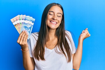 Young hispanic woman holding canadian dollars screaming proud, celebrating victory and success very...
