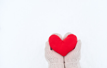 Obraz premium Creative greeting card for Valentines Day. Hold red soft heart toy made of fabric in beige mittens against background of white freshly fallen snow. Empty space for your greeting text.