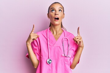 Young hispanic woman wearing doctor uniform and stethoscope amazed and surprised looking up and pointing with fingers and raised arms.