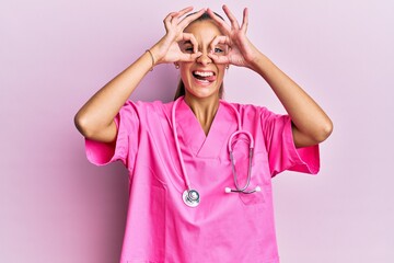 Young hispanic woman wearing doctor uniform and stethoscope doing ok gesture like binoculars...