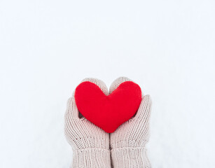 Fototapeta premium Creative greeting card for Valentines Day. Hold red soft heart toy made of fabric in beige mittens against background of white freshly fallen snow. Empty space for your greeting text.