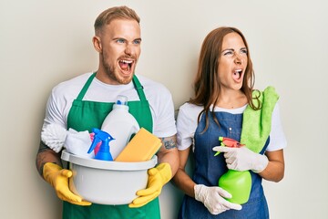 Young couple of girlfriend and boyfriend wearing apron holding products and cleaning spray angry and mad screaming frustrated and furious, shouting with anger. rage and aggressive concept.
