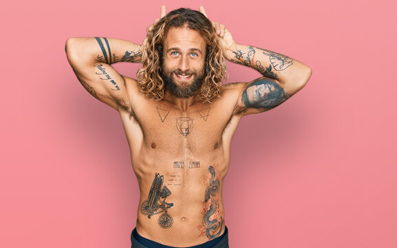 Handsome man with beard and long hair standing shirtless showing tattoos posing funny and crazy with fingers on head as bunny ears, smiling cheerful