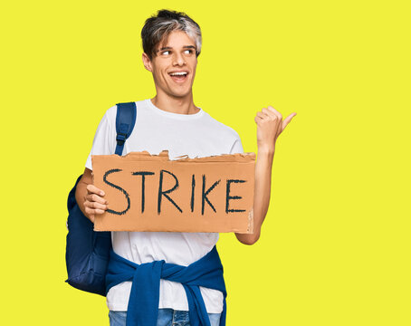 Young hispanic man holding strike banner cardboard pointing thumb up to the side smiling happy with open mouth