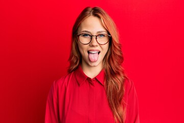 Young beautiful redhead woman wearing casual clothes and glasses over red background sticking tongue out happy with funny expression. emotion concept.