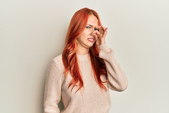 Young beautiful redhead woman wearing casual winter sweater smelling something stinky and disgusting, intolerable smell, holding breath with fingers on nose. bad smell