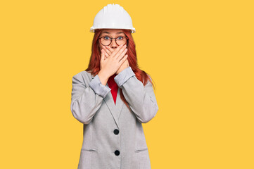 Young redhead woman wearing architect hardhat shocked covering mouth with hands for mistake. secret concept.