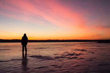 Fototapeta na wymiar The silhouette of a man standing on the ice of a frozen lake in the light of the sunset. Winter background