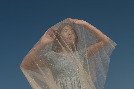 fineart portrait of a girl in blue dress holding thin tule veil with pearls under blue sky 
