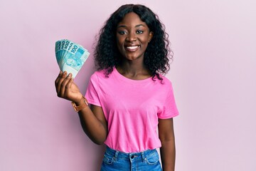 Beautiful african young woman holding 100 brazilian real banknotes looking positive and happy...