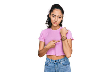 Hispanic teenager girl with dental braces wearing casual clothes in hurry pointing to watch time, impatience, looking at the camera with relaxed expression