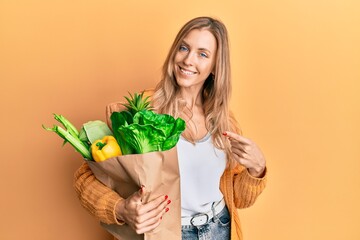 Beautiful caucasian woman holding paper bag with bread and groceries smiling happy pointing with hand and finger