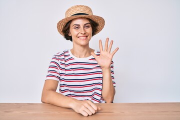 Young hispanic woman wearing casual clothes and hat sitting on the table showing and pointing up...