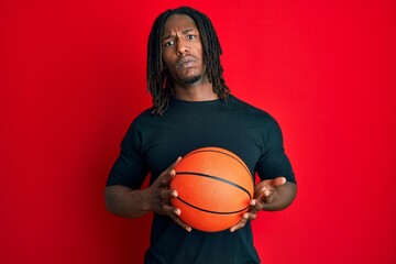 African american man with braids holding basketball ball clueless and confused expression. doubt concept.