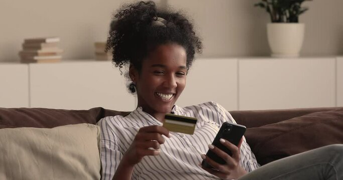 Young african woman rest on couch hold smartphone and credit card enjoy easy instant money transfer, makes good deed on-line donation secure payment. Happy customer and online retail services concept
